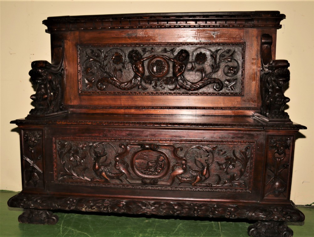 A dark carved walnut hall seat featuring high-relief carvings of mythical figures and floral scrollwork on the backrest and front storage panel. View XVII