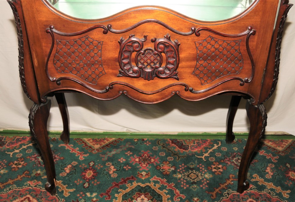 A dark brown carved mahogany display cabinet featuring glass doors and side panels, interior glass shelves, an ornate carved crest, and slender cabriole legs. View III