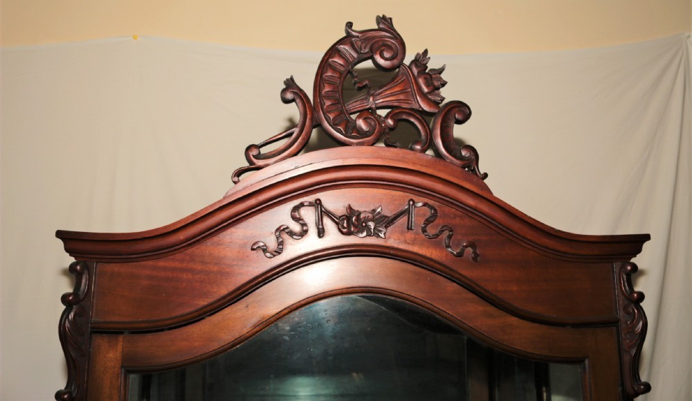 A dark brown carved mahogany display cabinet featuring glass doors and side panels, interior glass shelves, an ornate carved crest, and slender cabriole legs. View V