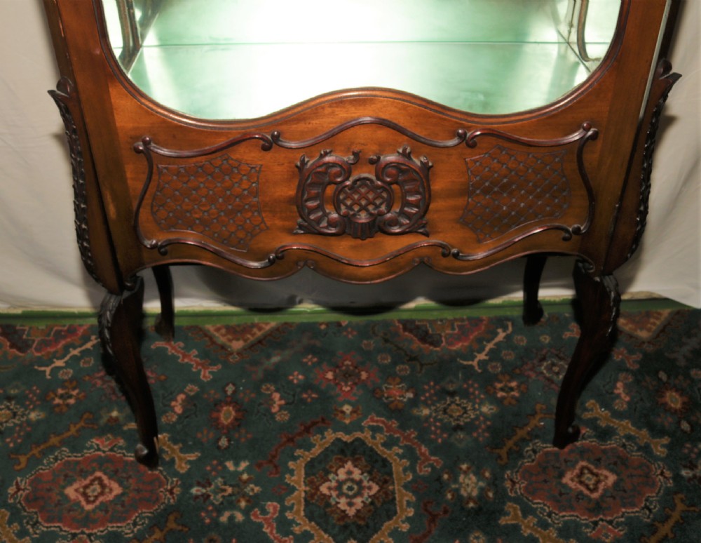 A dark brown carved mahogany display cabinet featuring glass doors and side panels, interior glass shelves, an ornate carved crest, and slender cabriole legs. View VI