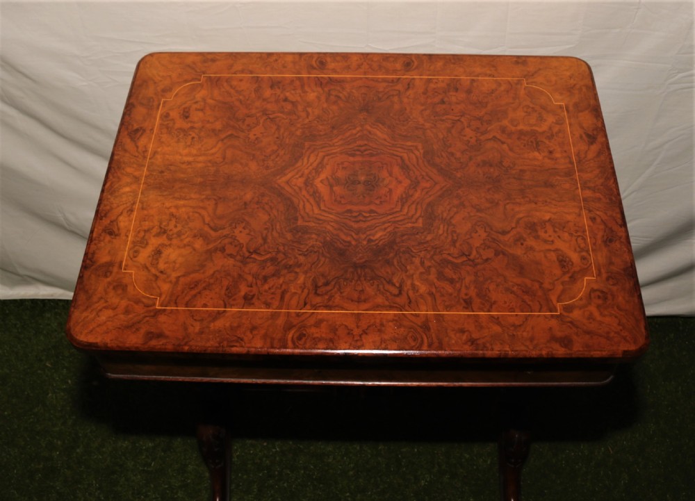 A Victorian burr walnut sewing table featuring a rectangular inlaid top, a hanging wooden work well, turned side supports, and carved legs on castors. View X