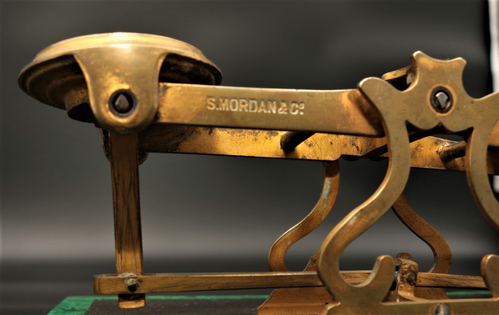 An antique brass postal balance scale set on a black stone base framed by a vibrant green malachite border. View III