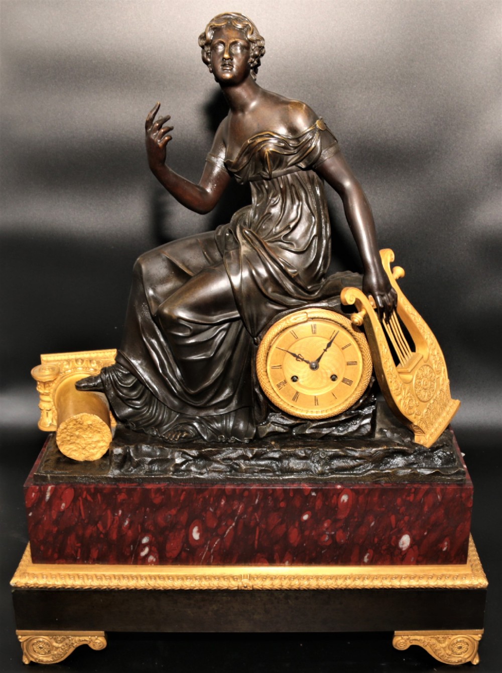 A French Empire mantel clock featuring a dark patinated bronze classical female figure holding a lyre, positioned beside a gold-coloured dial on a red marble base with gilt metal mounts.