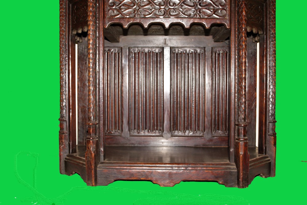 A dark carved oak Gothic Revival display cabinet featuring a glazed upper section, patterned fabric lining, and detailed linenfold panelling on the lower frame. View V