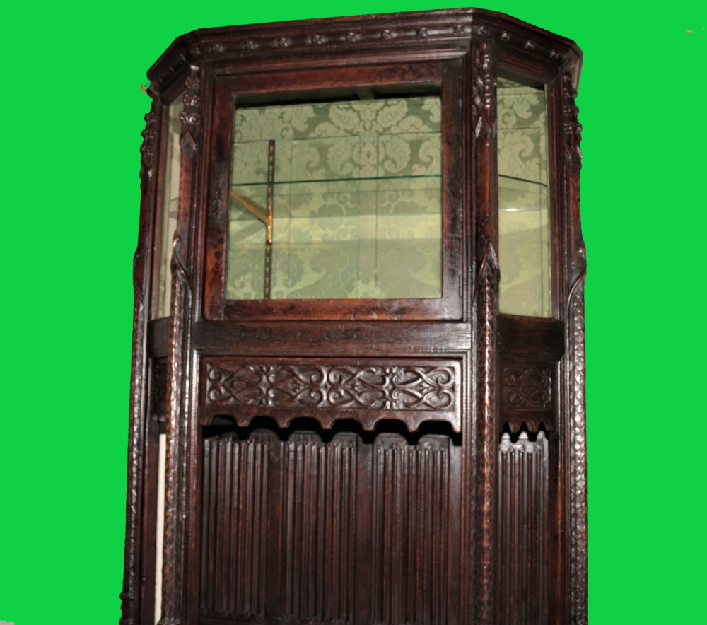 A dark carved oak Gothic Revival display cabinet featuring a glazed upper section, patterned fabric lining, and detailed linenfold panelling on the lower frame. View VII