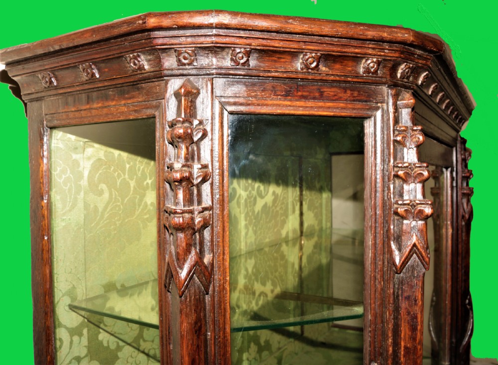 A dark carved oak Gothic Revival display cabinet featuring a glazed upper section, patterned fabric lining, and detailed linenfold panelling on the lower frame. View X