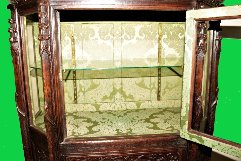 A dark carved oak Gothic Revival display cabinet featuring a glazed upper section, patterned fabric lining, and detailed linenfold panelling on the lower frame. View XI