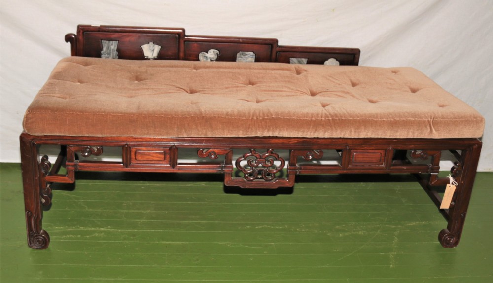 A carved dark hardwood Chinese daybed featuring a stepped backrest with marble panel inserts and a tufted light brown cushioned seat.