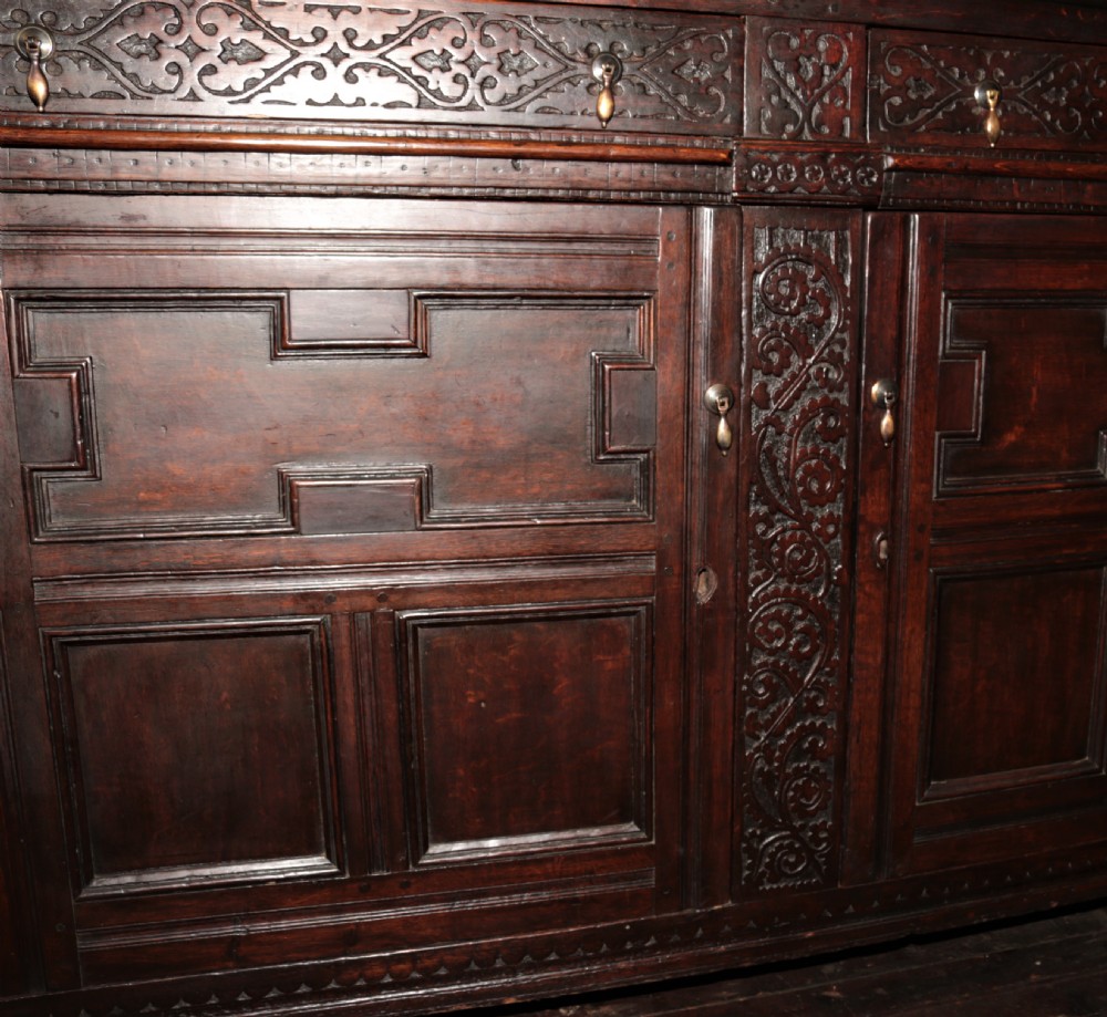 A dark carved oak Elizabethan court cupboard featuring geometric panelled doors, bulbous turned supports, and intricate foliate carvings. View XIII