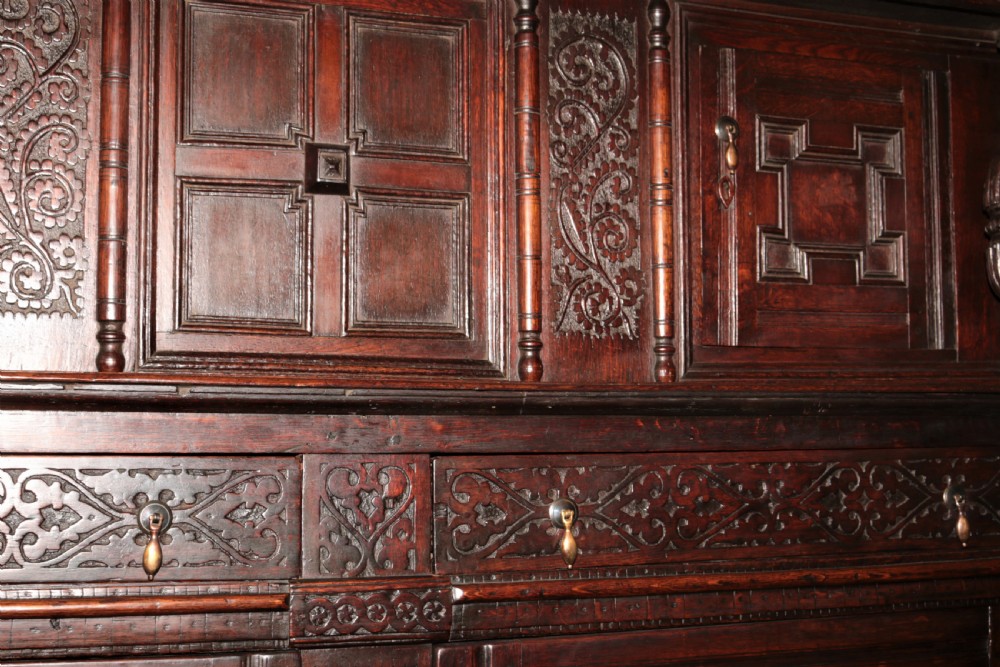 A dark carved oak Elizabethan court cupboard featuring geometric panelled doors, bulbous turned supports, and intricate foliate carvings. View XV