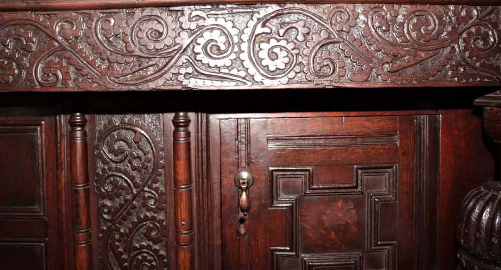 A dark carved oak Elizabethan court cupboard featuring geometric panelled doors, bulbous turned supports, and intricate foliate carvings. View XVII