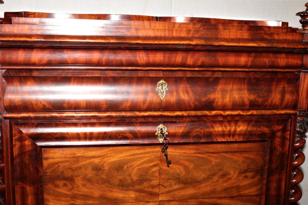 A Biedermeier flame mahogany fall-front bureau desk featuring twisted side columns, multiple drawers, brass keyhole escutcheons, and urn finials. View VI
