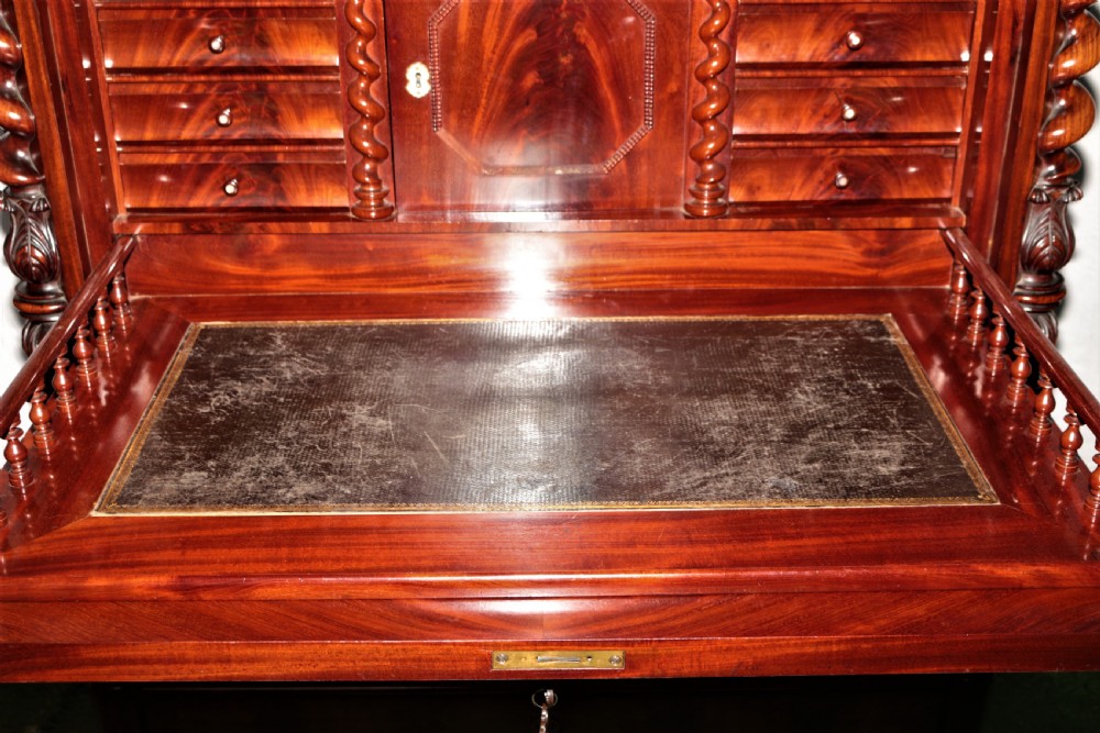 A Biedermeier flame mahogany fall-front bureau desk featuring twisted side columns, multiple drawers, brass keyhole escutcheons, and urn finials. View IX