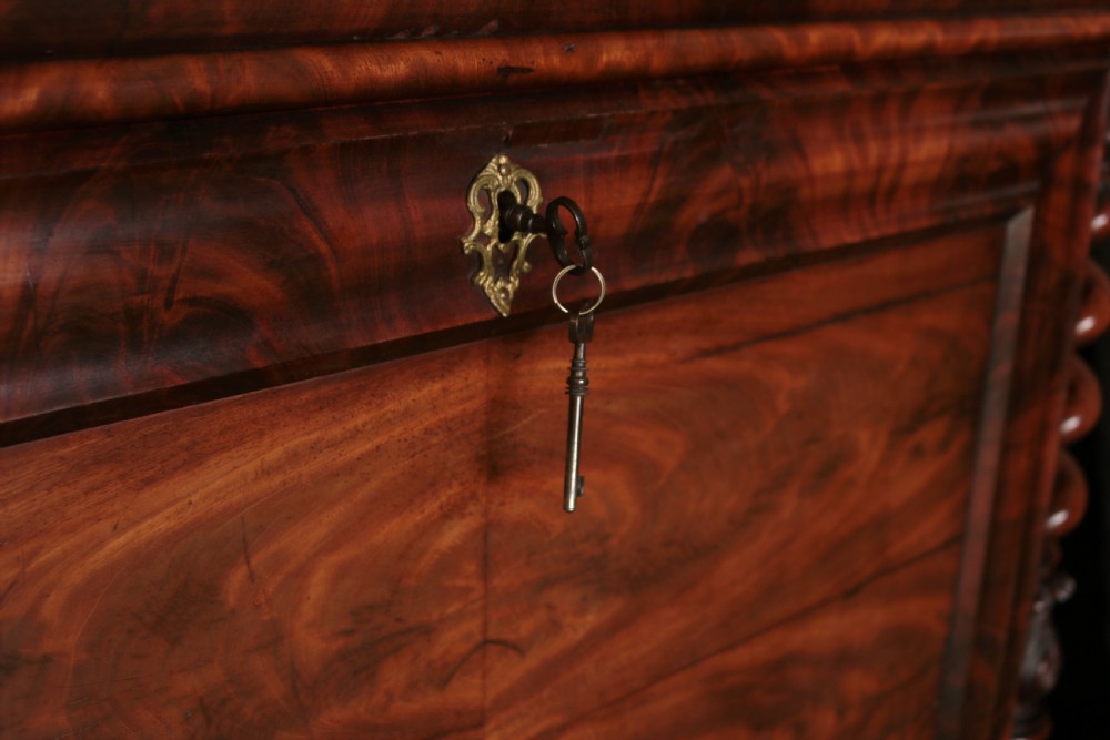 A Biedermeier flame mahogany fall-front bureau desk featuring twisted side columns, multiple drawers, brass keyhole escutcheons, and urn finials. View XV