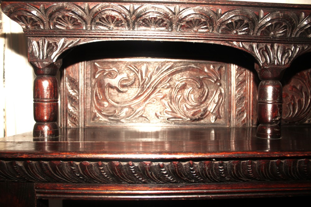 A dark carved oak dresser sideboard featuring intricate relief foliage panels, a central crest, upper shelving, and barley twist support columns. View III