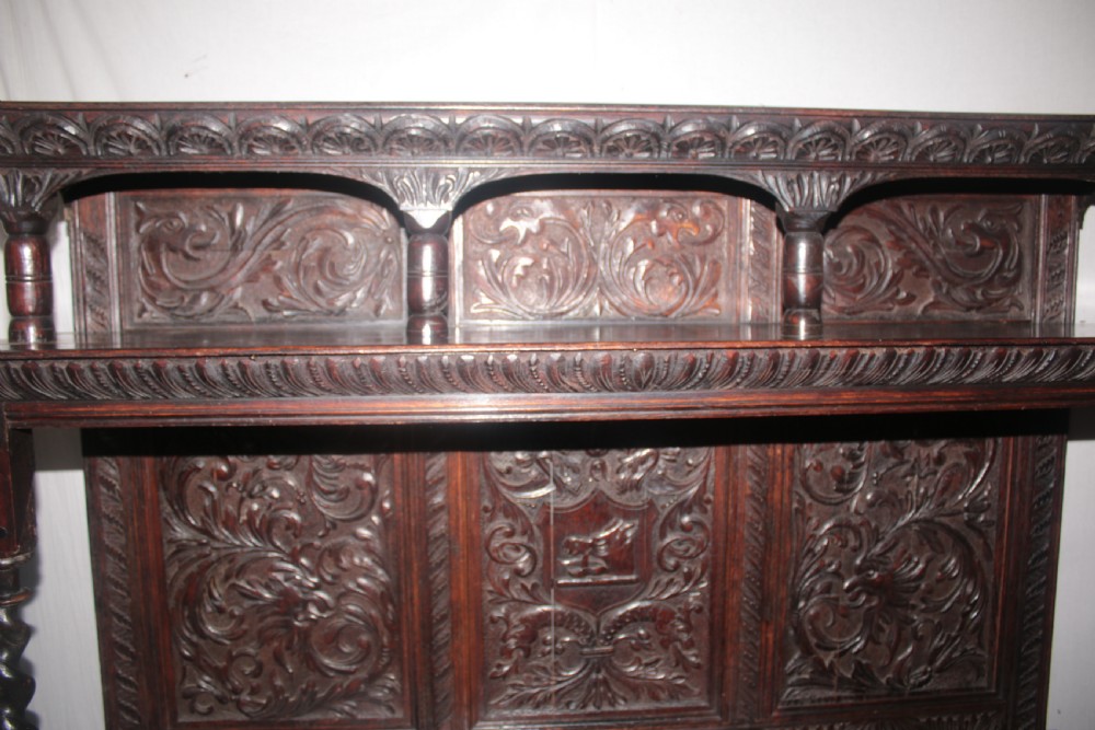 A dark carved oak dresser sideboard featuring intricate relief foliage panels, a central crest, upper shelving, and barley twist support columns. View IV