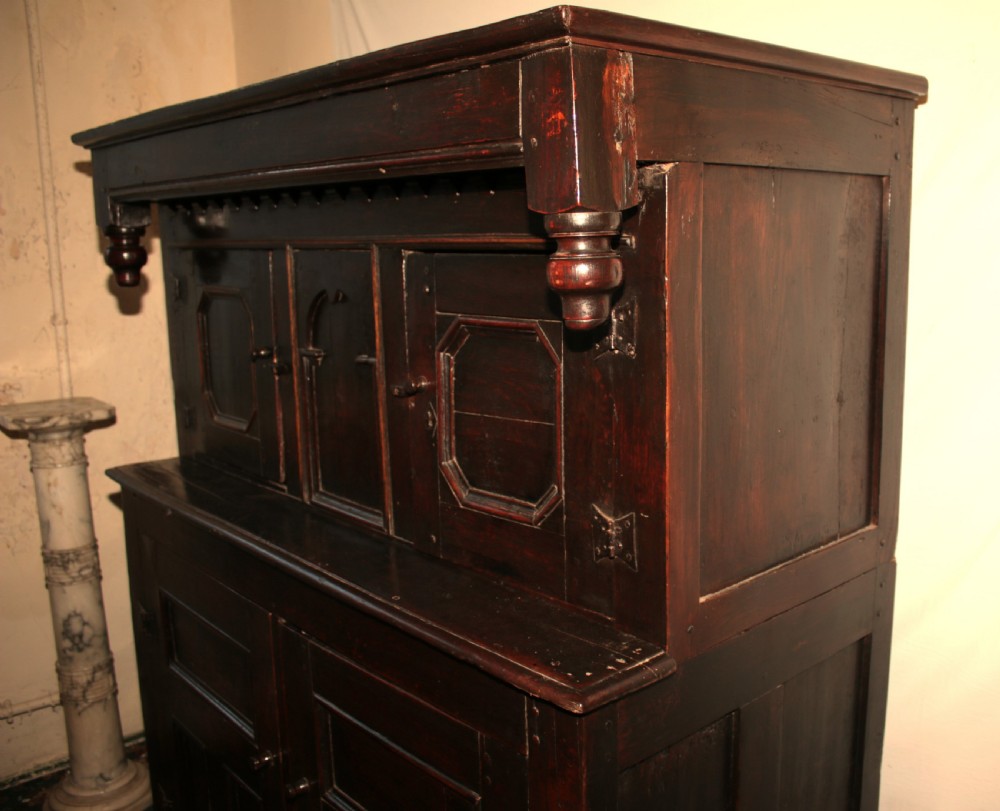 A dark oak court cupboard featuring a recessed upper section with carved panel doors and drop pendants above a lower two-door cabinet with iron hinges. View III
