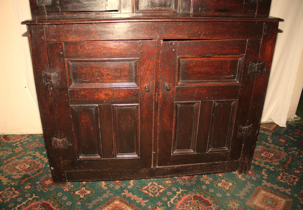 A dark oak court cupboard featuring a recessed upper section with carved panel doors and drop pendants above a lower two-door cabinet with iron hinges. View VI