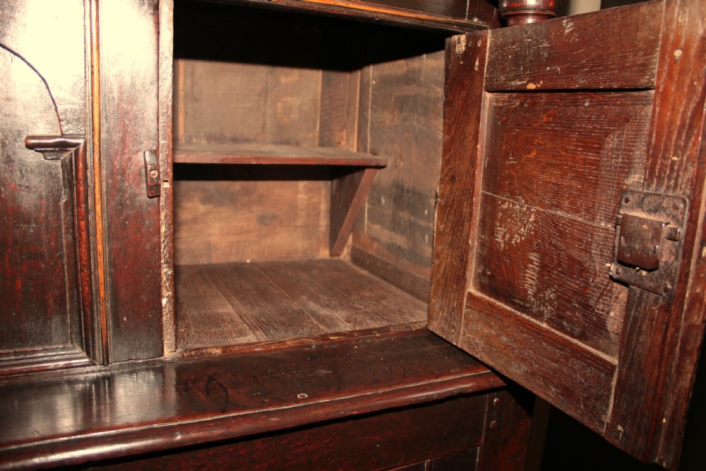 A dark oak court cupboard featuring a recessed upper section with carved panel doors and drop pendants above a lower two-door cabinet with iron hinges. View VIII