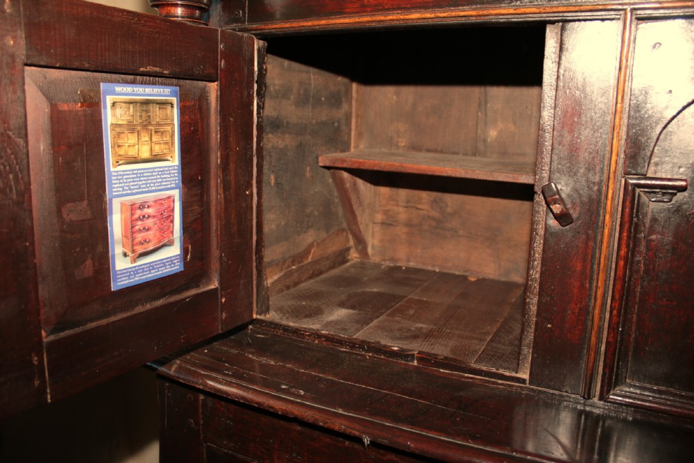 A dark oak court cupboard featuring a recessed upper section with carved panel doors and drop pendants above a lower two-door cabinet with iron hinges. View IX