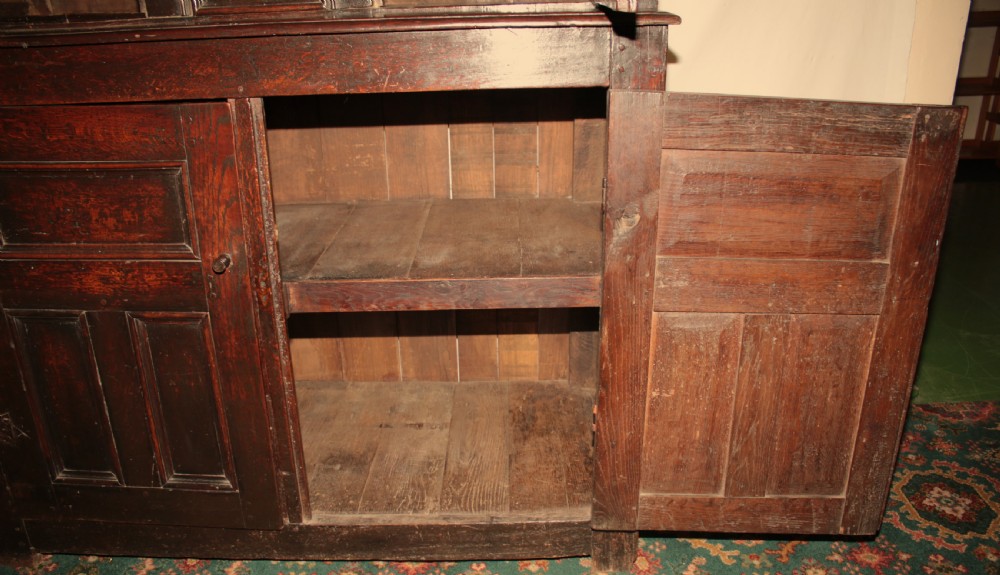 A dark oak court cupboard featuring a recessed upper section with carved panel doors and drop pendants above a lower two-door cabinet with iron hinges. View X