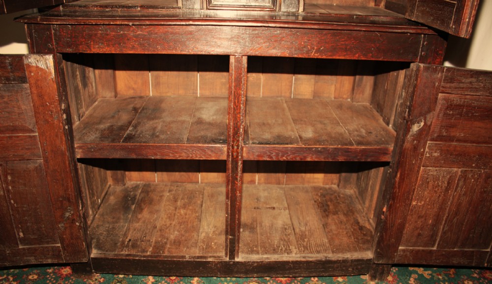 A dark oak court cupboard featuring a recessed upper section with carved panel doors and drop pendants above a lower two-door cabinet with iron hinges. View XII