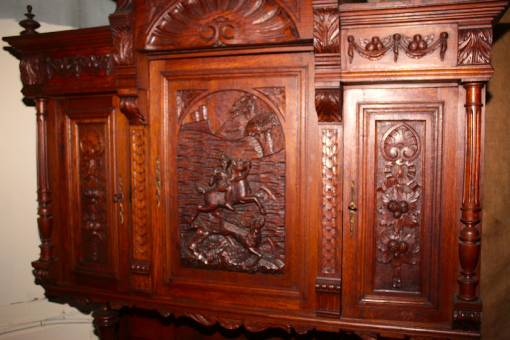 A large, two-tiered dark oak cabinet featuring intricate relief carvings, a central panel depicting a hunting scene, turned columns, and carved lower cupboards. View III