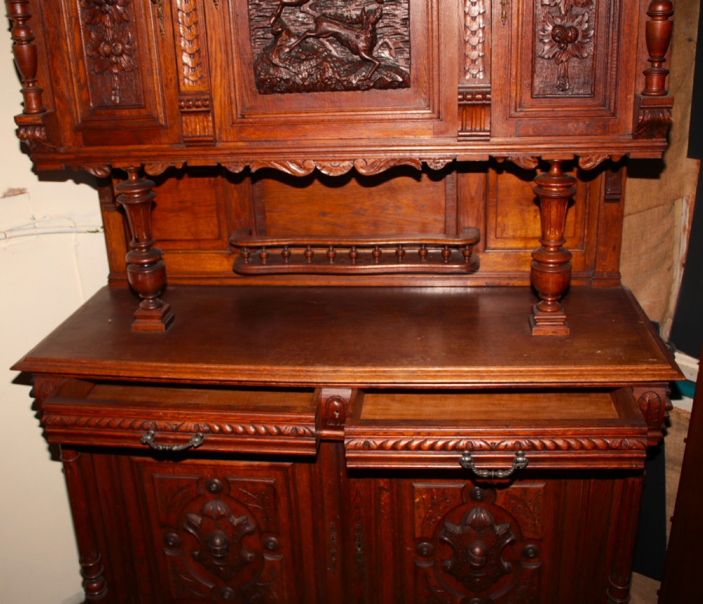 A large, two-tiered dark oak cabinet featuring intricate relief carvings, a central panel depicting a hunting scene, turned columns, and carved lower cupboards. View IV