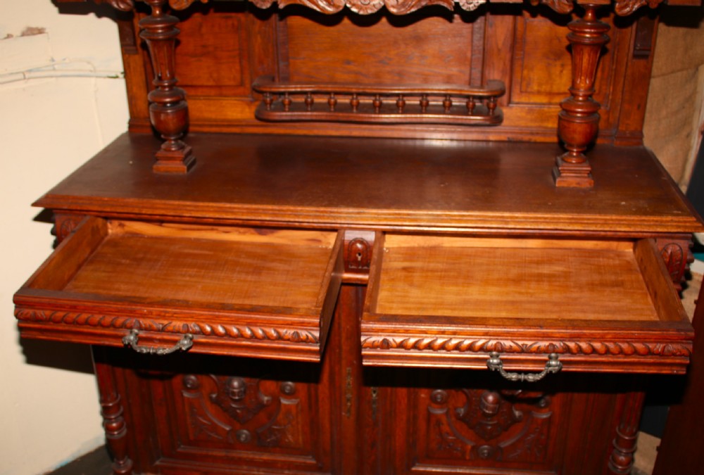 A large, two-tiered dark oak cabinet featuring intricate relief carvings, a central panel depicting a hunting scene, turned columns, and carved lower cupboards. View V