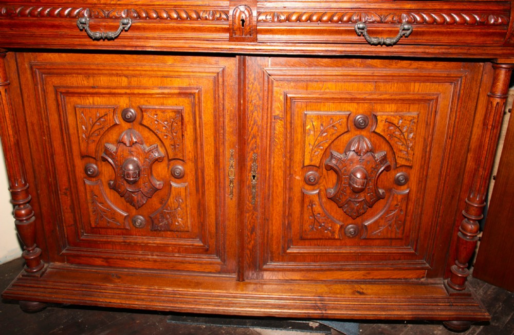 A large, two-tiered dark oak cabinet featuring intricate relief carvings, a central panel depicting a hunting scene, turned columns, and carved lower cupboards. View IX