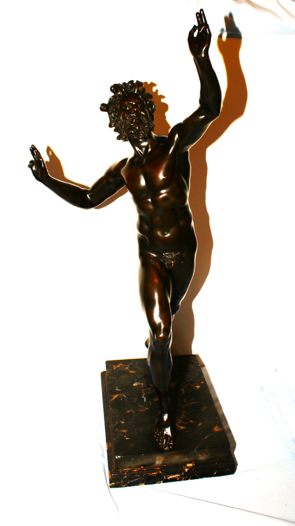 A dark patinated bronze sculpture of a dancing faun with raised arms mounted on a square marble base.