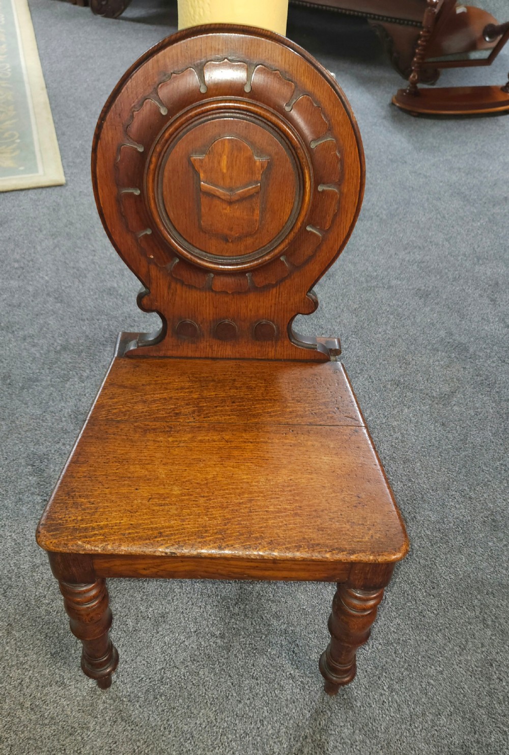 A Victorian oak hall chair featuring a rounded carved backrest with a central shield motif, a solid flat wooden seat, and turned front legs.
