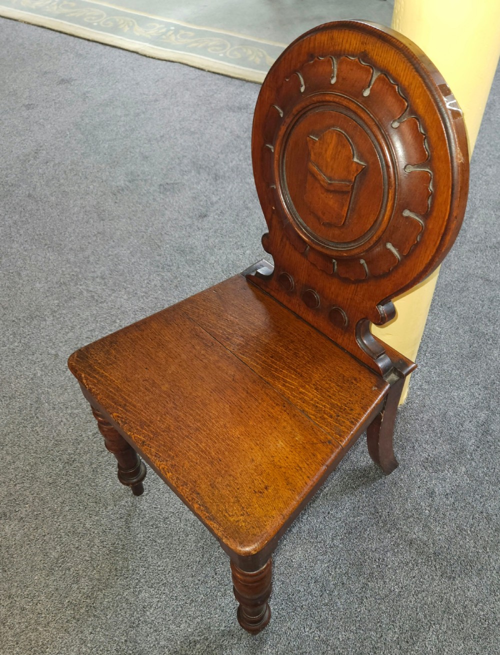 A Victorian oak hall chair featuring a rounded carved backrest with a central shield motif, a solid flat wooden seat, and turned front legs. View IV