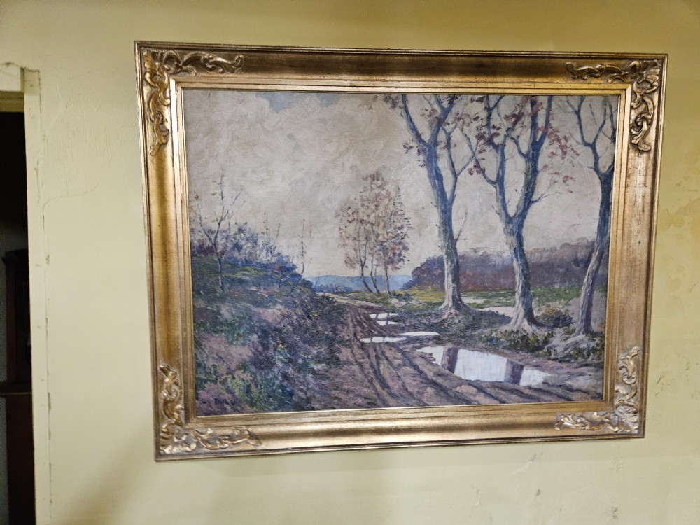A large framed oil painting depicting a muddy countryside path lined with bare trees under an overcast sky, housed in an ornate gilt frame.