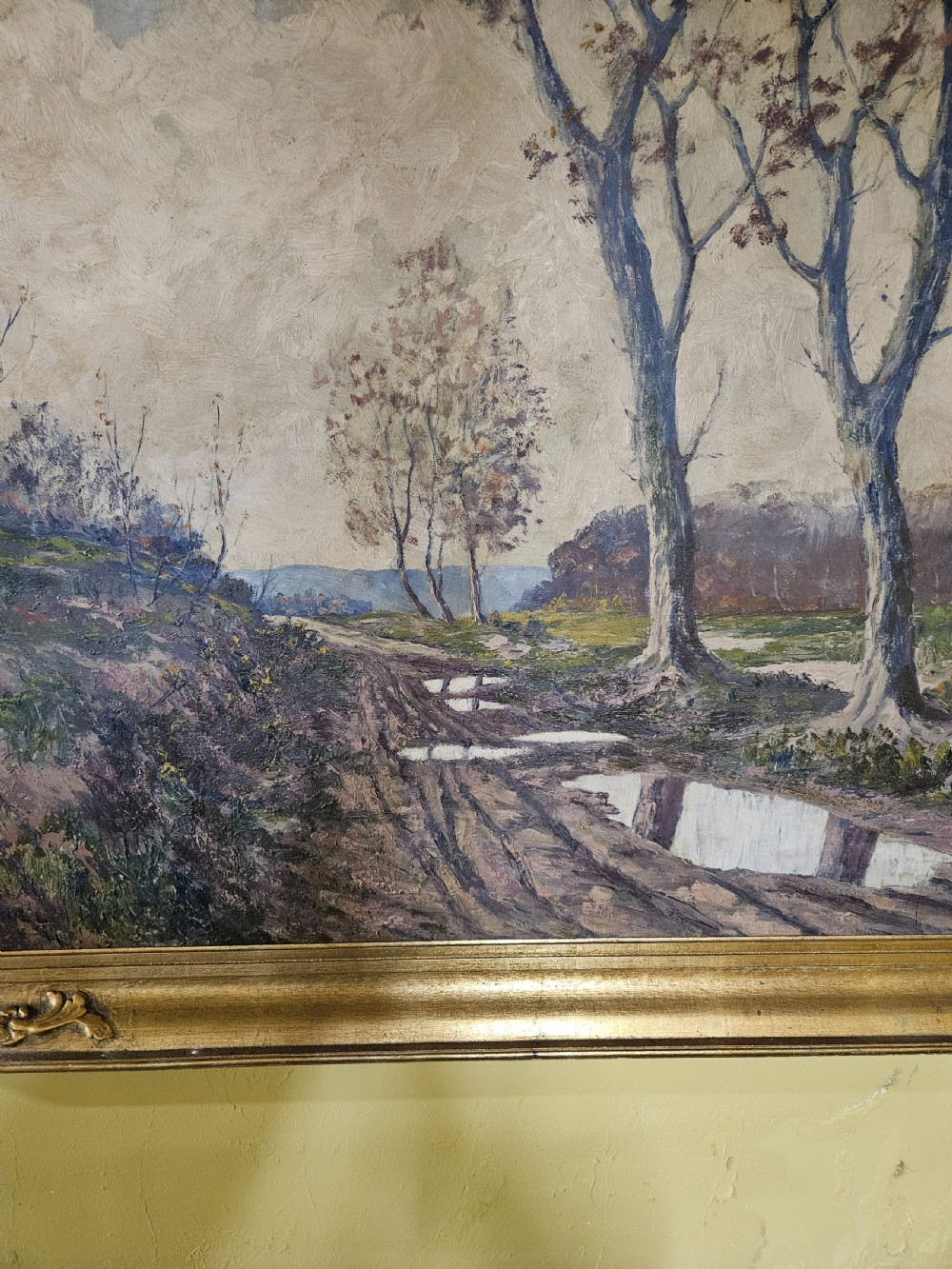 A large framed oil painting depicting a muddy countryside path lined with bare trees under an overcast sky, housed in an ornate gilt frame. View II