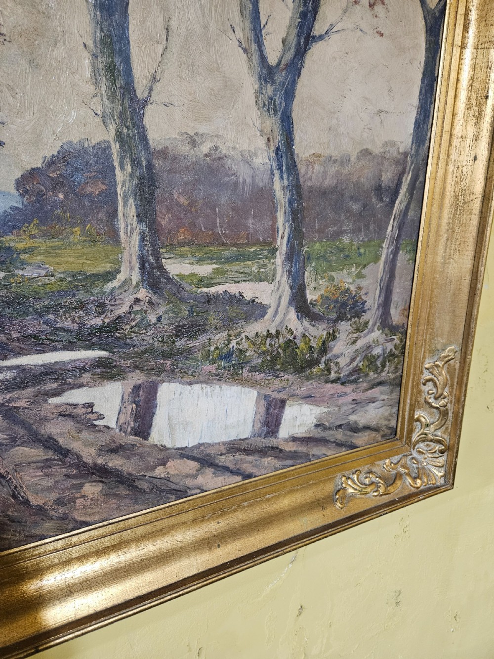 A large framed oil painting depicting a muddy countryside path lined with bare trees under an overcast sky, housed in an ornate gilt frame. View IV