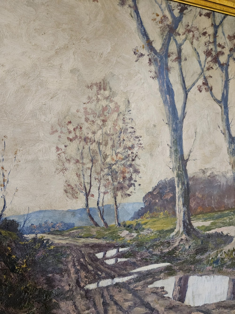 A large framed oil painting depicting a muddy countryside path lined with bare trees under an overcast sky, housed in an ornate gilt frame. View V