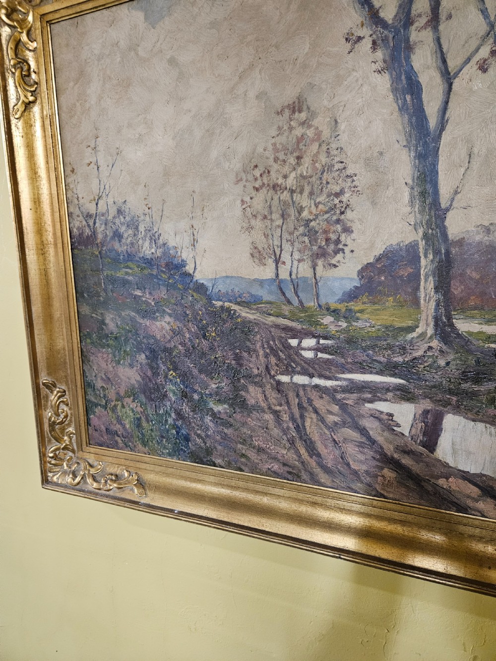 A large framed oil painting depicting a muddy countryside path lined with bare trees under an overcast sky, housed in an ornate gilt frame. View VI