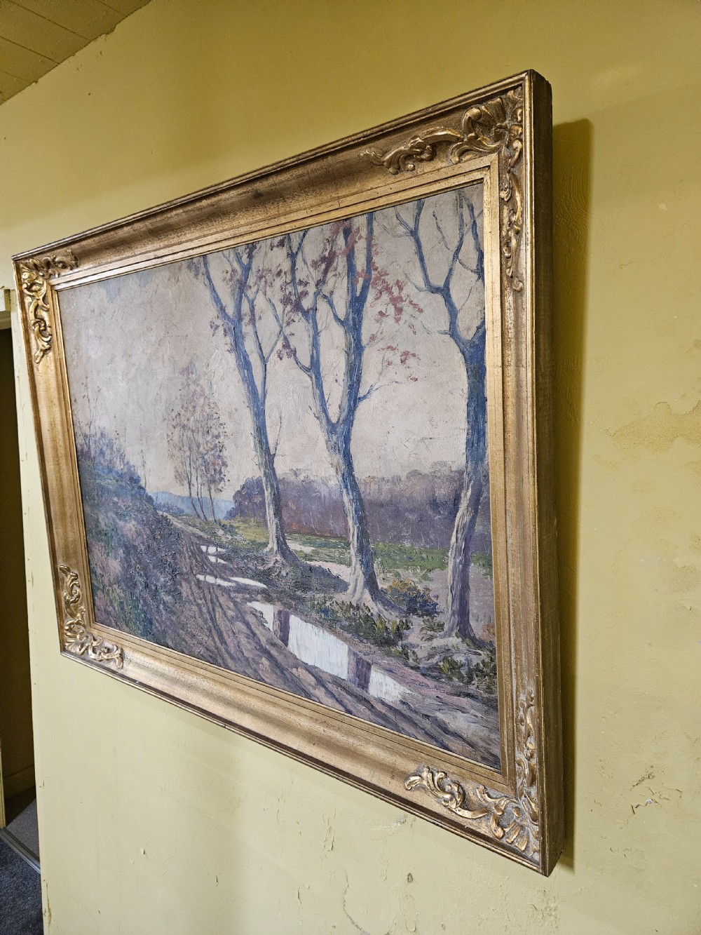 A large framed oil painting depicting a muddy countryside path lined with bare trees under an overcast sky, housed in an ornate gilt frame. View VII