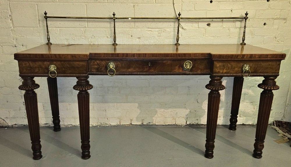 A Regency mahogany serving table featuring a brass gallery rail, lion mask ring handles, and six reeded tapered legs.