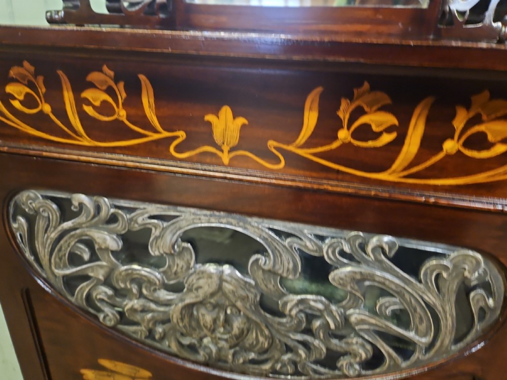 An Art Nouveau dark mahogany music cabinet featuring detailed wood marquetry, a pierced metal cartouche panel, and an upper mirrored gallery shelf. View X