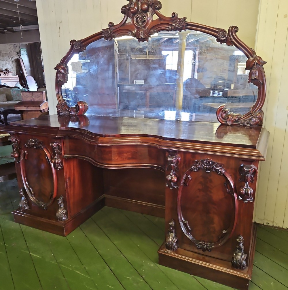 A Victorian mahogany sideboard with a large carved scrollwork framed mirror top, a serpentine surface, and twin pedestal cupboards with oval mouldings.