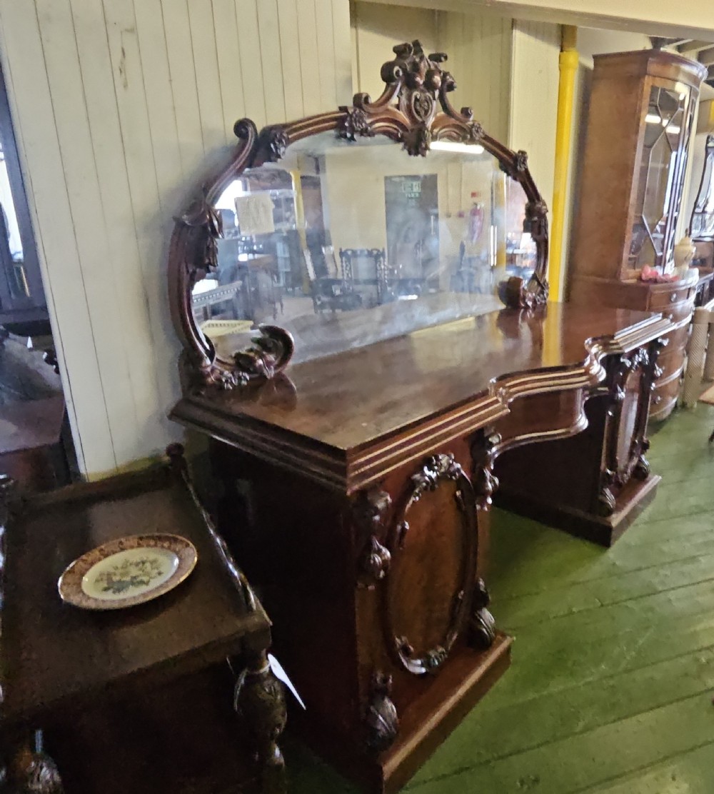A Victorian mahogany sideboard with a large carved scrollwork framed mirror top, a serpentine surface, and twin pedestal cupboards with oval mouldings. View IX