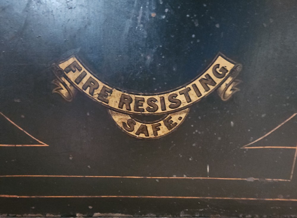 A black Victorian cast iron safe featuring a central brass makers plaque, gold line decoration, a brass T-bar handle, and a key inserted into the lock. View VI