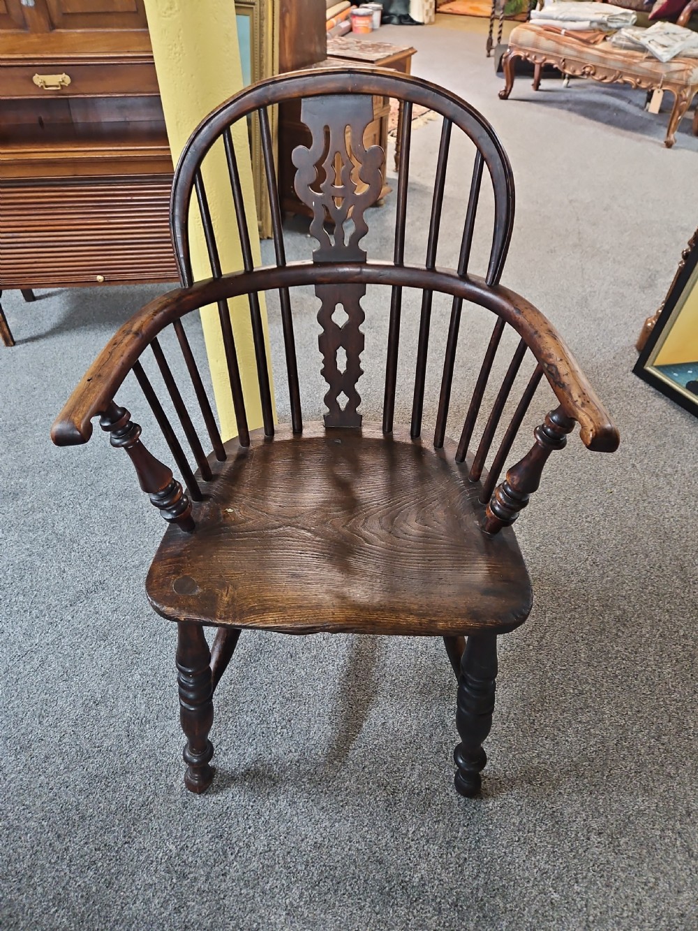 An antique dark yew wood Windsor armchair featuring a curved hoop back, a pierced central splat, turned spindles, and carved wooden arms supported by turned legs.