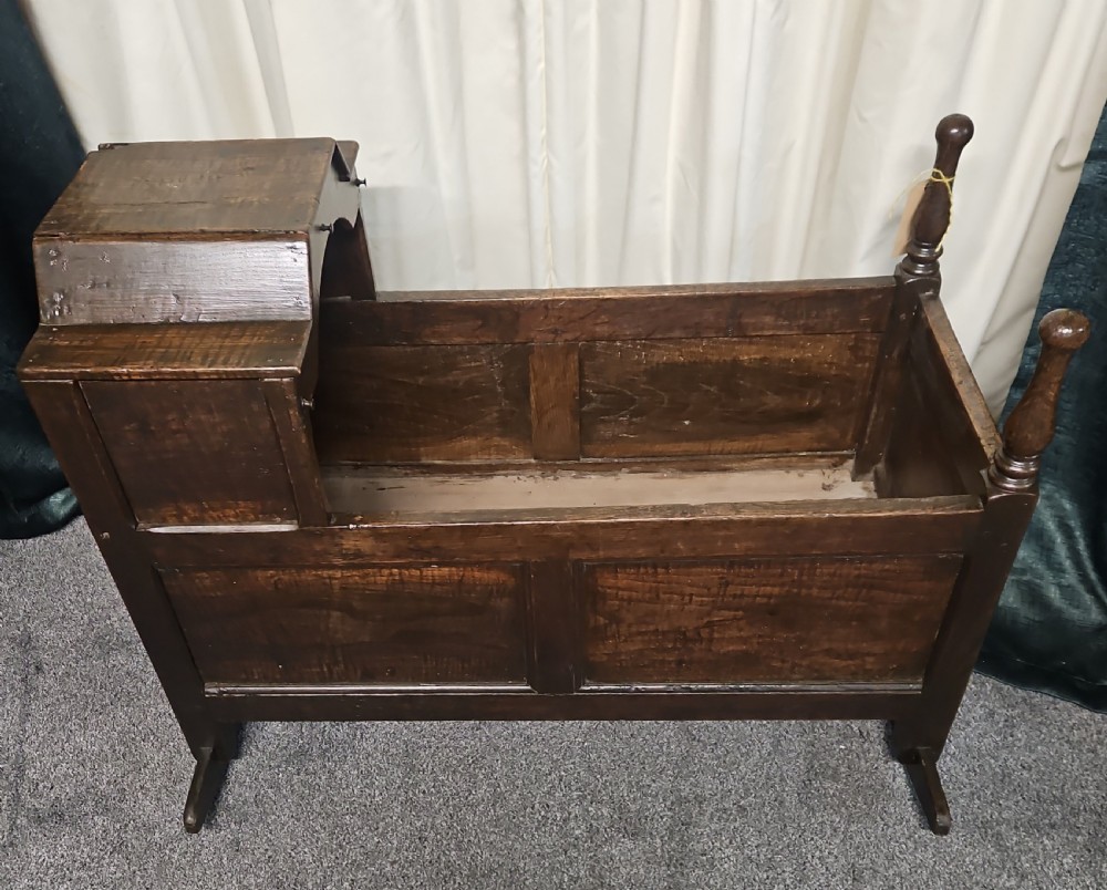 A dark oak antique rocking cradle featuring a panelled frame, an enclosed hood at one end, turned corner posts, and wooden rockers.