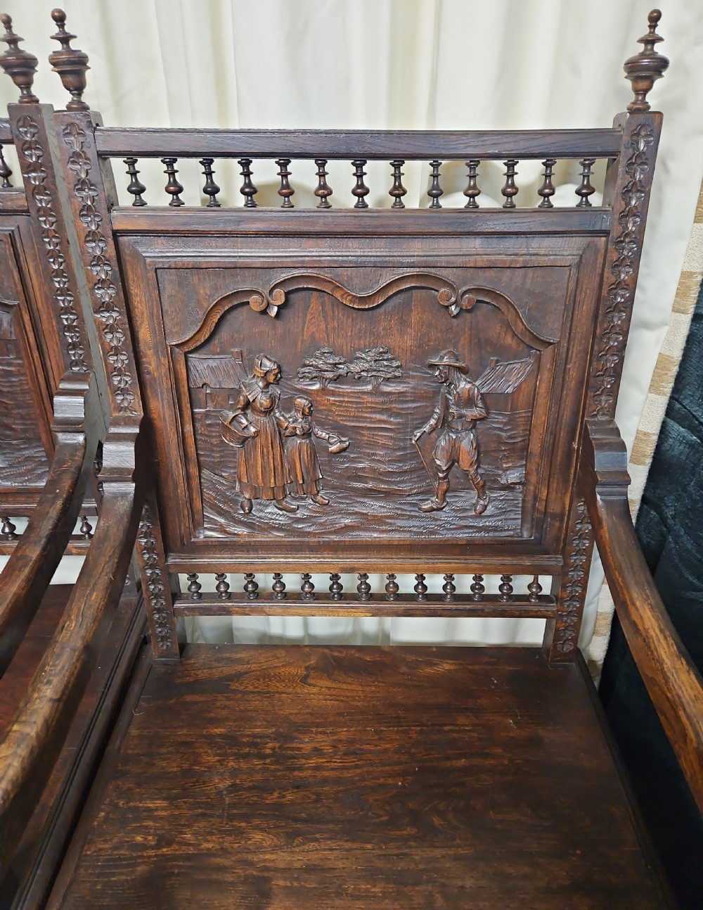 A pair of dark carved oak Breton armchairs featuring backrest panels with relief scenes of folk figures and turned spindle galleries. View XII