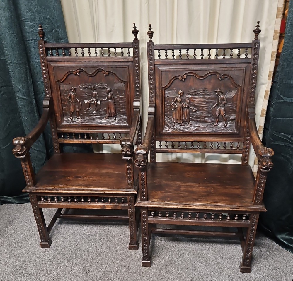 A pair of dark carved oak Breton armchairs featuring backrest panels with relief scenes of folk figures and turned spindle galleries.