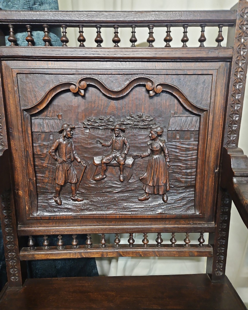 A pair of dark carved oak Breton armchairs featuring backrest panels with relief scenes of folk figures and turned spindle galleries. View VI