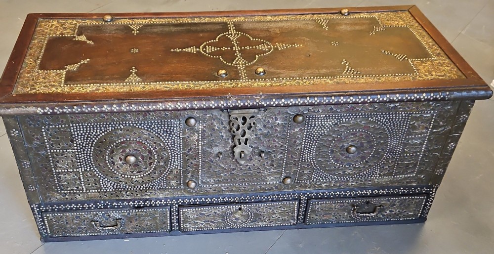 A mid-Victorian wooden Zanzibar chest decorated with intricate brass studs, geometric brass sheeting, and three base drawers.